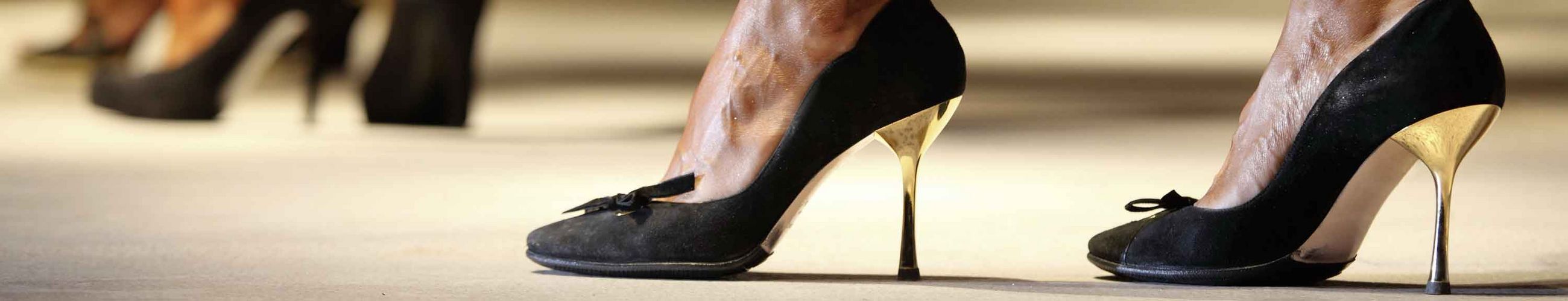 Das Bild zeigt die Füße einer Frau in hohen, schwarzen Schuhen, im Hintergrund sieht man verschwommen weitere.